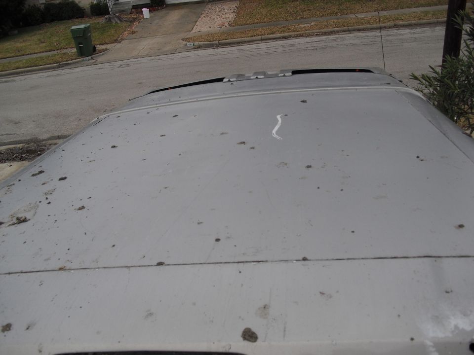 Top view of a dusty vehicle roof with some debris on it. A street and sidewalk are visible in the background