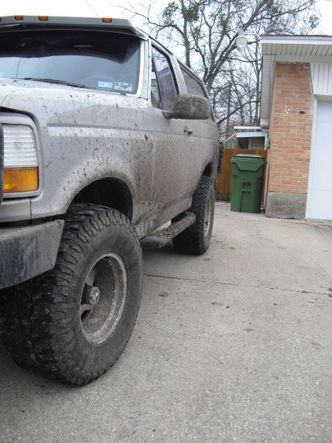 1992 Ford Bronco - A muddy 1992 Ford Bronco is parked next to a brick garage and a green trash bin.
