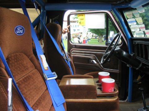 1986 Ford Bronco - The interior of a 1986 Ford Bronco features brown velour seats, blue racing harnesses, and a blue roll cage.