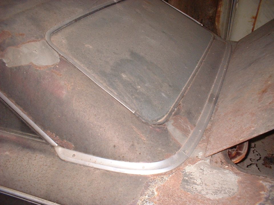 Close-up of a rusty car roof with a metal trim around a window opening. A wheel is visible on the right