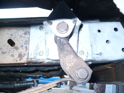 1979 Ford Bronco - Close-up under a truck frame rail showing a fabricated bare-steel bracket plate welded and bolted to the rusty boxed frame, carrying a pivo…