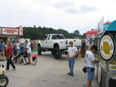 1986 Ford Bronco - A lifted white pickup truck is parked at an outdoor event with people milling around food vendors.