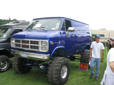 1986 Ford Bronco - A lifted blue GMC van with massive tires is displayed at an outdoor event, with another vehicle visible to the left.