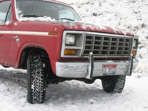 1981 Ford Bronco - A red Ford Bronco is parked in the snow, showing its front grille and off-road tires.