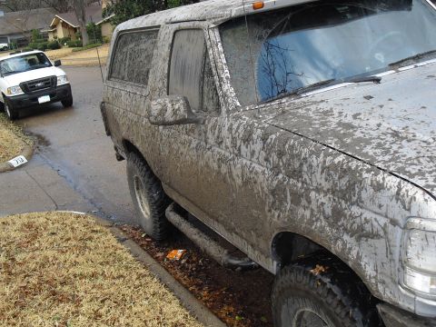 1992 Ford Bronco - A heavily mud-splattered 1992 Ford Bronco is parked on the side of a street, with another vehicle visible in the background.