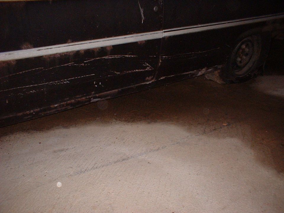 Side view of a dark colored car with a white stripe, showing rust and damage near the bottom, with a tire visible