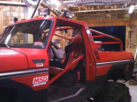 1979 Ford Bronco - Red 1979 Ford Bronco with doors and top removed, showing a bright red roll cage, black-and-red racing bucket seats, roof-mounted light bars…