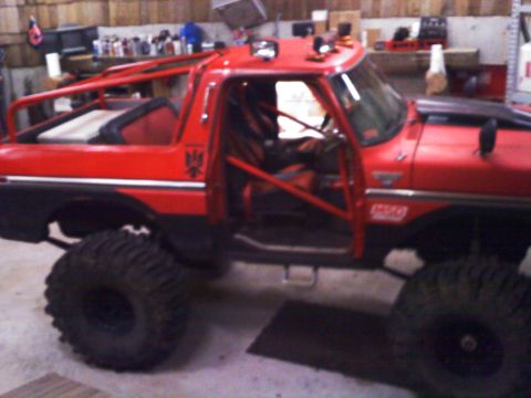 1979 Ford Bronco - Side view of a lifted red full-size Ford Bronco in a shop, top and doors removed, with a red-painted exterior roll cage extending over the…