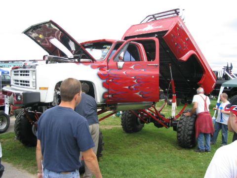 1986 Ford Bronco - This heavily modified pickup truck features custom flame paint, an open hood, and a raised bed, displayed at an outdoor event.
