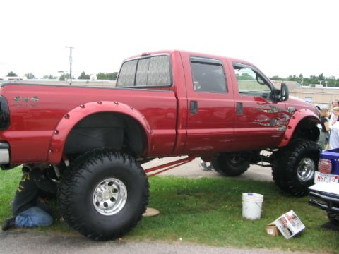1986 Ford Bronco - A red lifted pickup truck is parked on grass, with a person working underneath it.