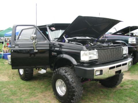 1986 Ford Bronco - A black Ford Bronco is displayed with its hood open at a truck show, with another vehicle visible behind it.