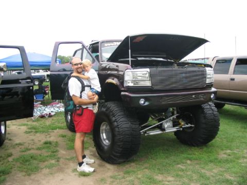 1986 Ford Bronco - A lifted Ford pickup truck with its hood open is displayed at an outdoor event, with a man and child standing nearby.
