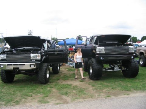1986 Ford Bronco - Two lifted black Ford trucks are displayed with their hoods open at a truck show, with one identified as a 1986 Ford Bronco.