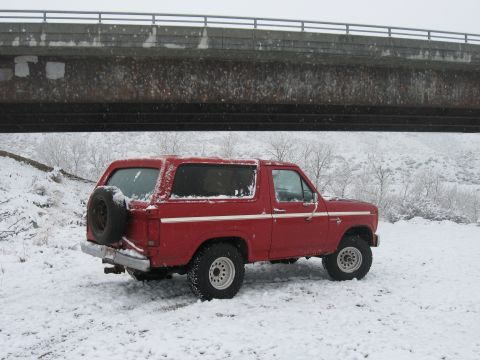 1981 Ford Bronco - A red Ford Bronco sits in the snow, viewed from the rear three-quarter angle under an overpass.