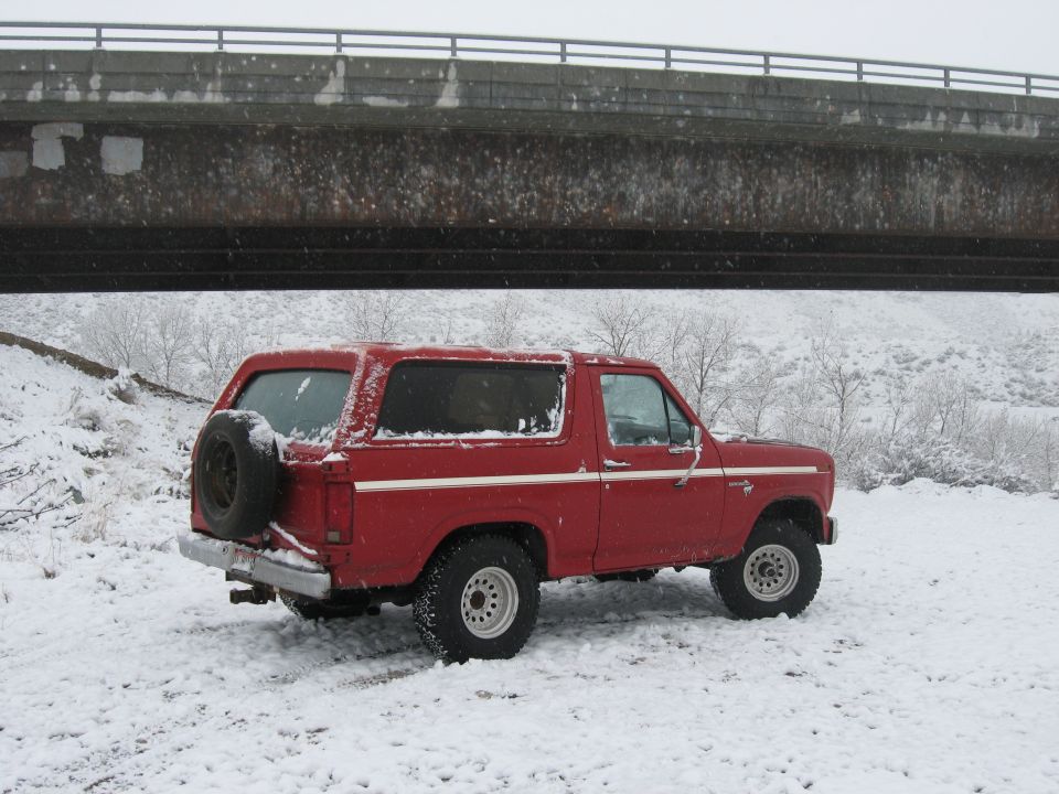 Red Ford Bronco with spare tire on rear, parked in snow under a bridge