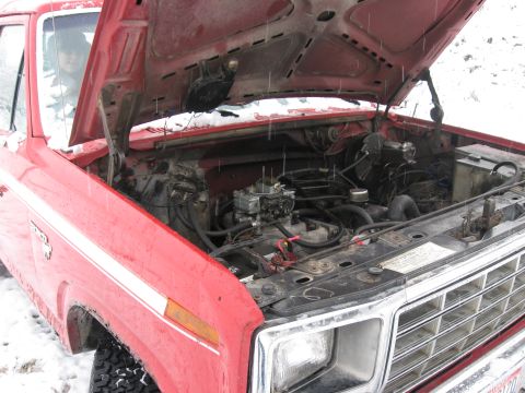 1981 Ford Bronco - Engine bay of a red 1980s Ford Bronco/F-Series with the hood raised, showing a V8 with a carburetor, air cleaner removed, and various vacuu…