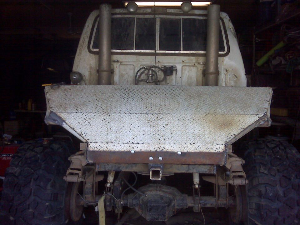 Rear view of a white pickup truck with huge tires, dual exhaust stacks, and a diamond plate spreader attachment
