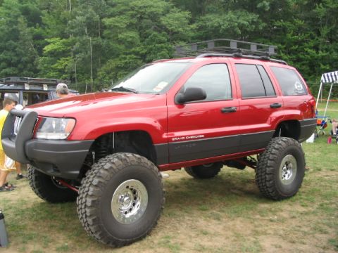 1986 Ford Bronco - A lifted red Jeep Grand Cherokee is parked on grass with trees in the background.