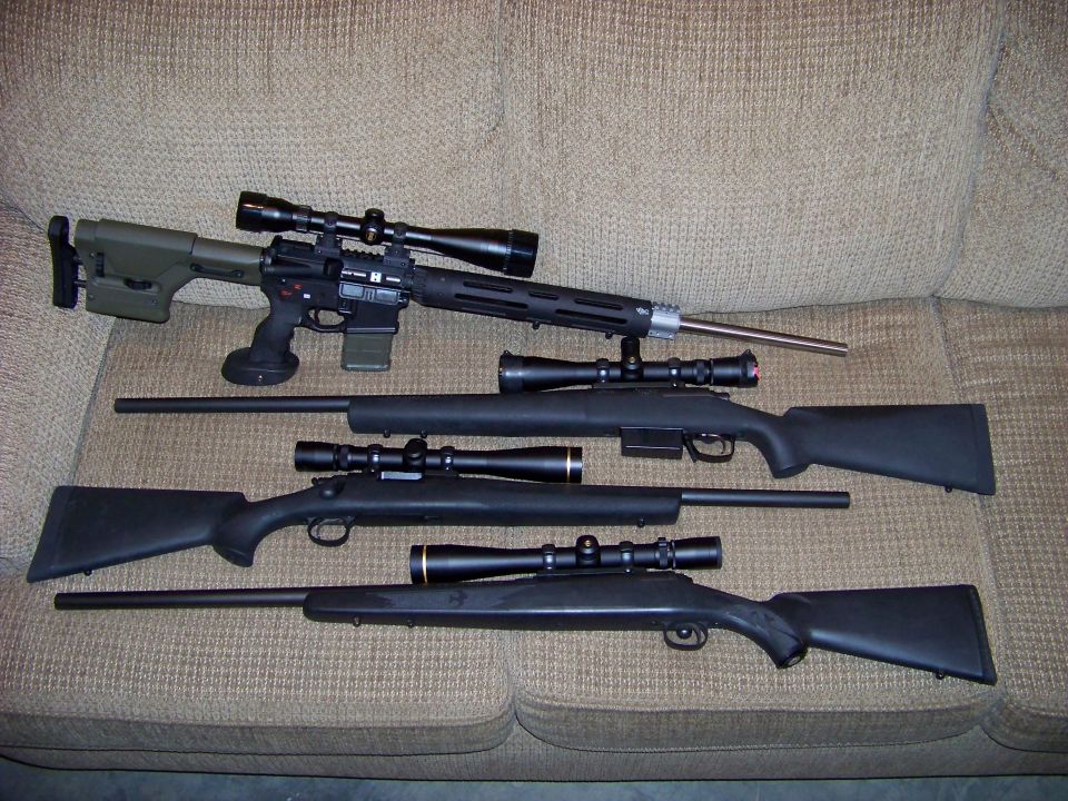Four scoped rifles laid out on a tan couch, including an AR-pattern and three bolt-actions