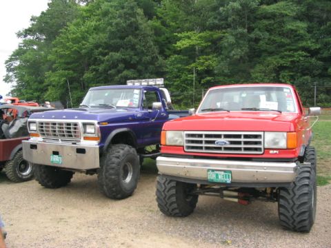 1986 Ford Bronco - Two lifted Ford trucks, a blue one and a red one, are parked side-by-side at an outdoor show.