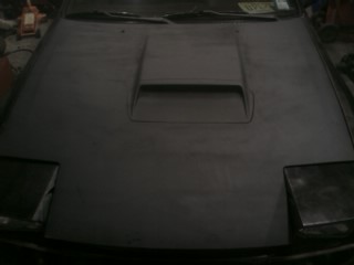1987 Mazda RX-7 - Front view of a car hood with a prominent air intake and pop-up headlights, showing a Supermotors logo.