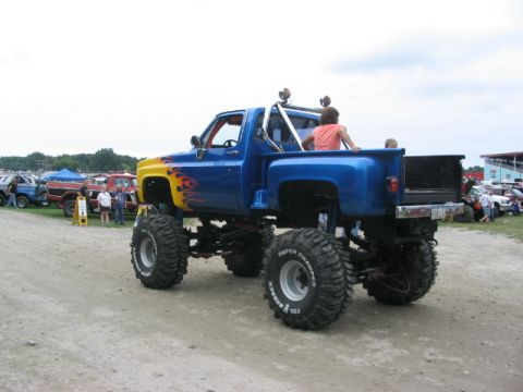 1986 Ford Bronco - A lifted pickup truck with flame decals is parked at an outdoor event with other vehicles in the background.