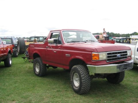 1986 Ford Bronco - A lifted red Ford Bronco is parked on a grassy field with other vehicles and people in attendance.