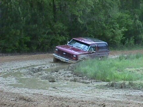 1984 Ford Bronco - This maroon Ford Bronco is stuck in a muddy rut, with trees lining the background.