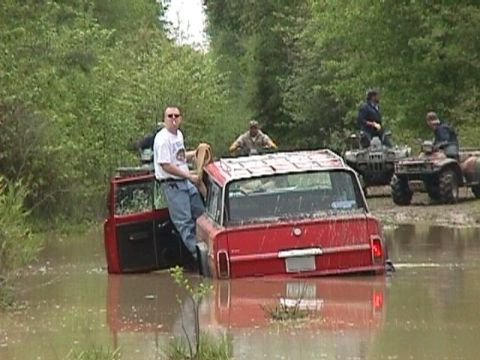 1984 Ford Bronco - A red Jeep is submerged in muddy water with people around and ATVs in the background.