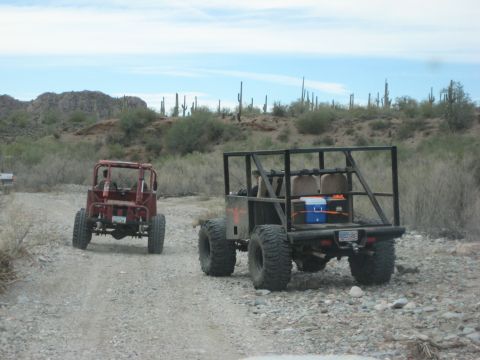 1996 Ford Bronco - Two custom off-road vehicles navigate a dusty desert trail under a bright blue sky with saguaro cacti in the background.