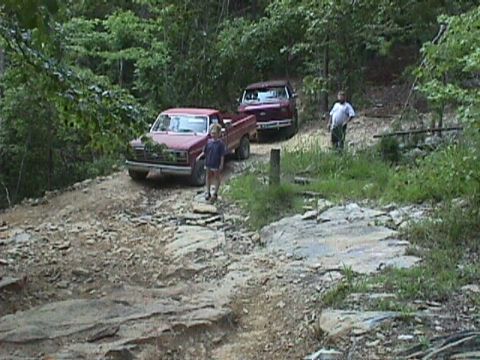 1984 Ford Bronco - Two pickup trucks navigate a rough, off-road trail with trees and rocks visible.