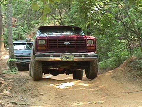 1984 Ford Bronco - A maroon Ford Bronco navigates a dirt path in a wooded area, followed closely by another vehicle.