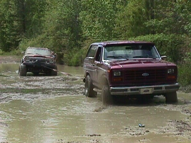 Maroon 1980 Ford Bronco driving through mud, with a Jeep in the background