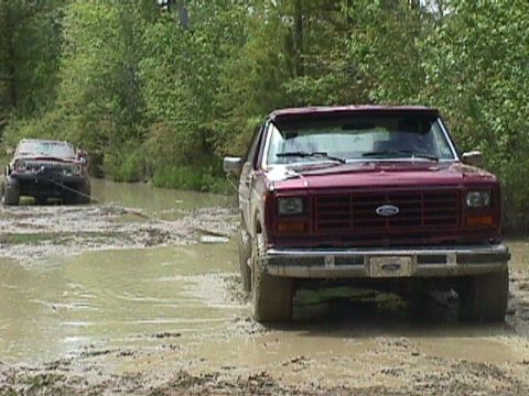 1984 Ford Bronco - Two off-road vehicles navigate a muddy trail with trees in the background.