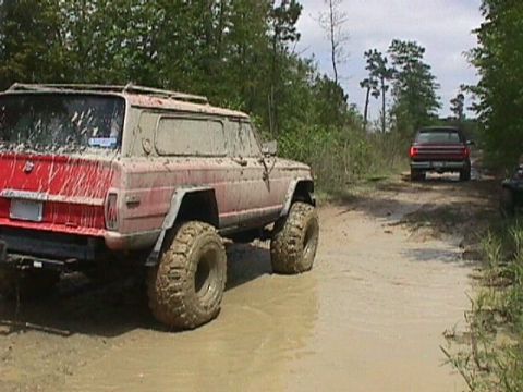 1984 Ford Bronco - A red SUV navigates a muddy off-road trail with another vehicle in the distance.