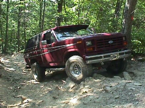1984 Ford Bronco - Maroon Ford Bronco navigating a rocky, wooded trail. The vehicle appears to be from the early 1980s.