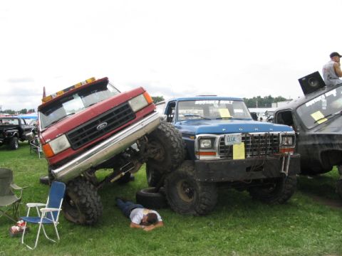 1986 Ford Bronco - A man naps on the grass as two heavily modified Ford trucks are displayed at an outdoor event.