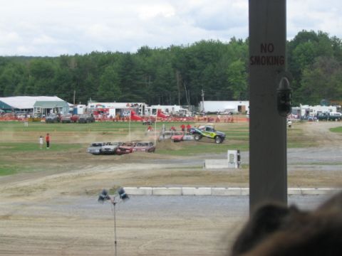 1986 Ford Bronco - A monster truck is airborne over a pile of cars at a truck show, with spectators and buildings in the background.
