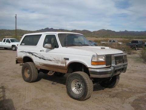 1996 Ford Bronco - This lifted white Ford Bronco is parked in a dusty, outdoor area with other vehicles visible in the distance.