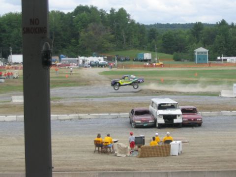 1986 Ford Bronco - A car is captured mid-air as it jumps a dirt ramp during an event, with spectators watching.