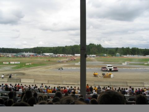 1986 Ford Bronco - Spectators watch an event unfold at a fairground or stadium with multiple buildings and a treeline visible.