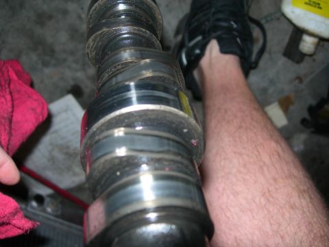 1984 Ford Bronco - A camshaft is shown close-up, with a hand and a red cloth on the left and a person's leg and shoe on the right.
