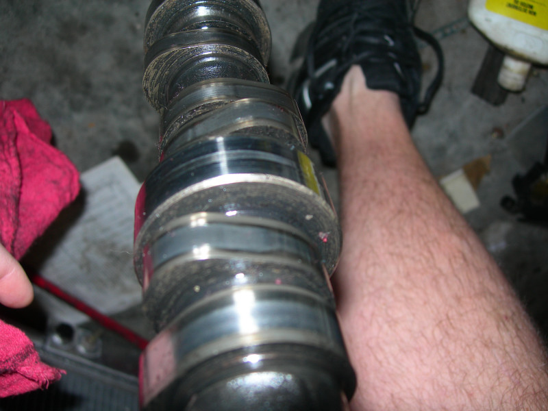 Camshaft with dirt and oil residue, held by a hand with a red cloth, next to a person's leg and shoe