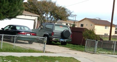 1986 Ford Bronco - A gray Ford Bronco is parked in a driveway next to a black sedan, with houses in the background.