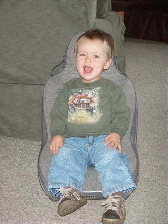 Toddler sitting in a car seat, wearing a green shirt with a truck graphic and blue jeans
