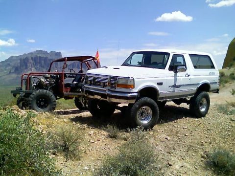 1996 Ford Bronco - A white Ford Bronco and a custom off-road buggy sit parked on a dusty trail under a bright blue sky.