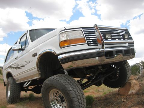 1996 Ford Bronco - A white 1996 Ford Bronco is shown from a low front-left angle, lifted and positioned on rocks with a chrome grille and aftermarket wheels.