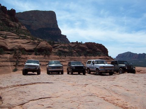 1996 Ford Bronco - A fleet of SUVs, including a white 1996 Ford Bronco, are parked on a rocky desert terrain under a partly cloudy sky.