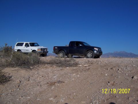 1996 Ford Bronco - Two trucks, a 1996 Ford Bronco and a black pickup, are parked on a dusty hill with mountains in the distance.