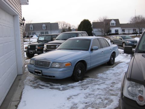 1999 Ford Crown Victoria - Light blue sedan parked in a snowy driveway with other vehicles present.